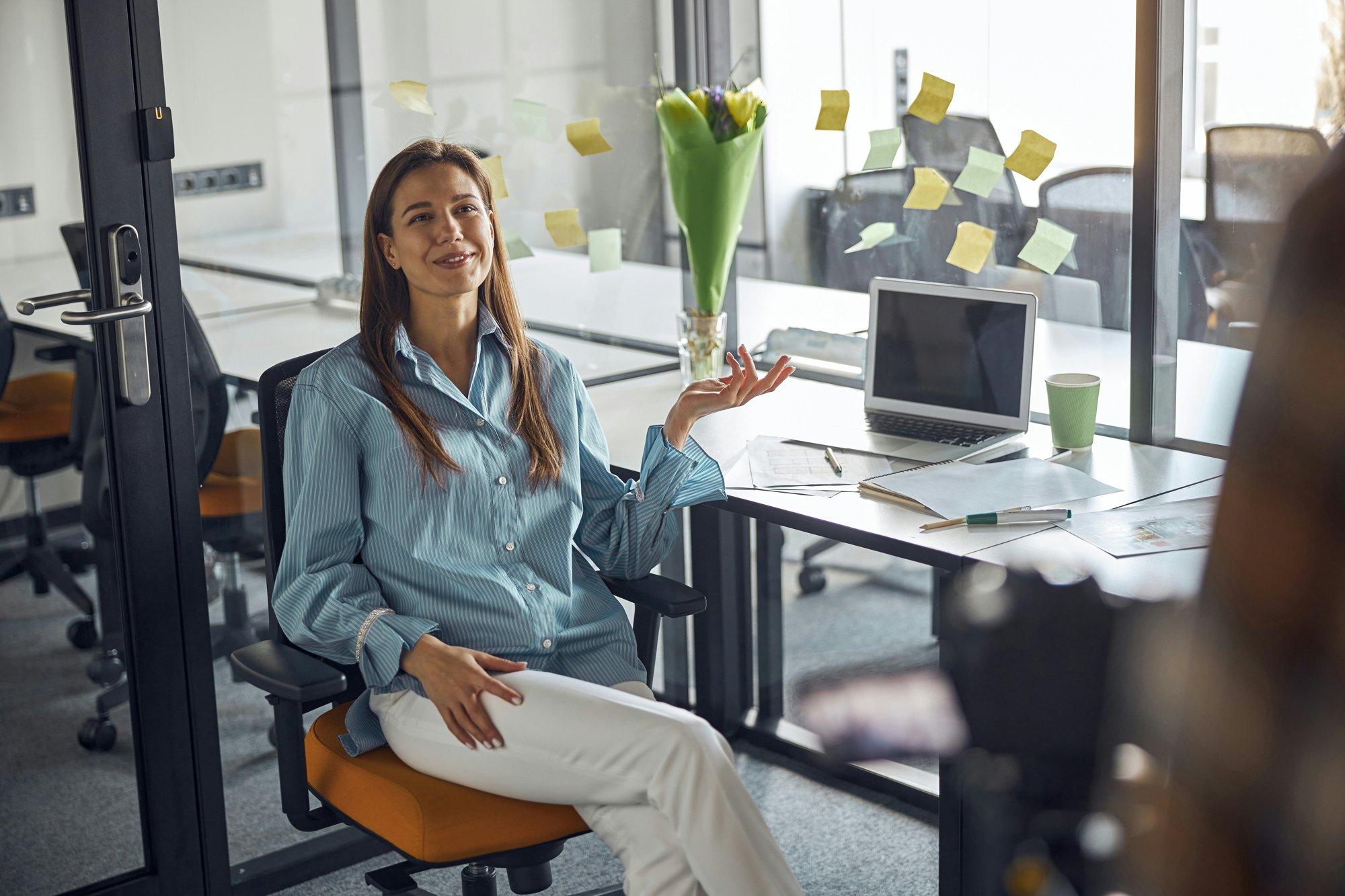 Smiling woman in a striped blue shirt sits at an office desk talking with a colleague, sticky notes on the glass wall behind her.