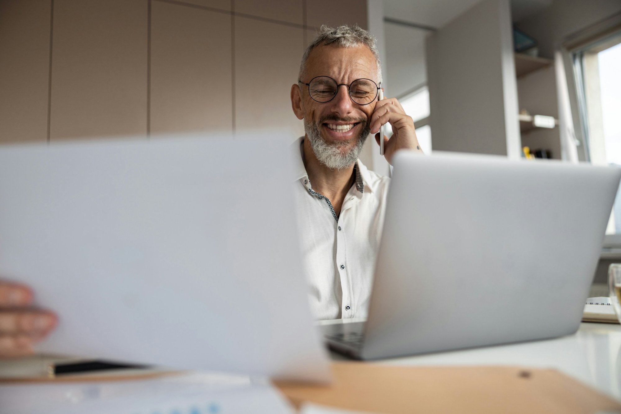 Smiling grey-haired man with glasses and a beard talks on the phone at his desk beside an open laptop.